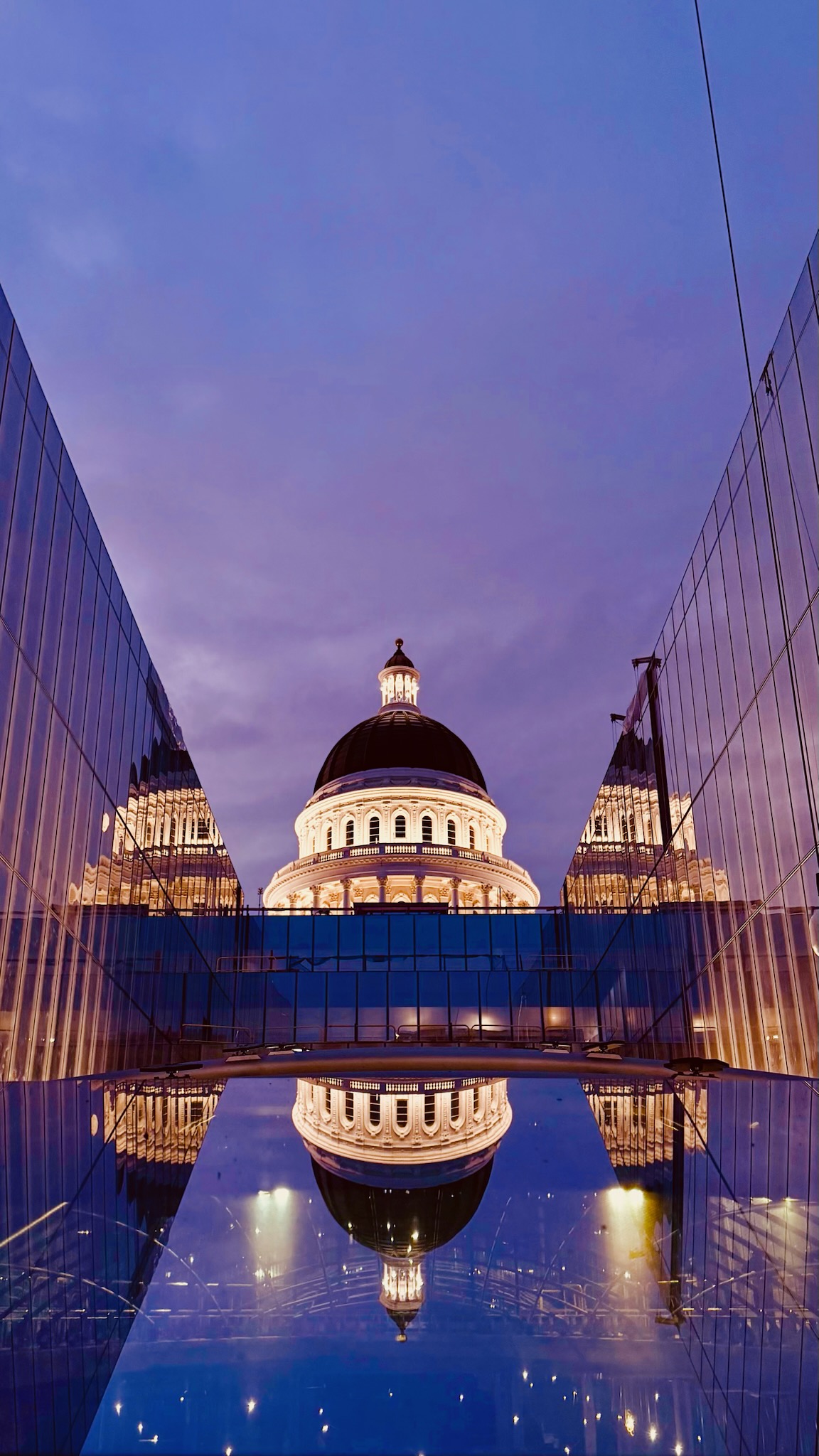 Vertical view of the illuminated Capitol Dome reflected in the glass Annex roof at twilight.