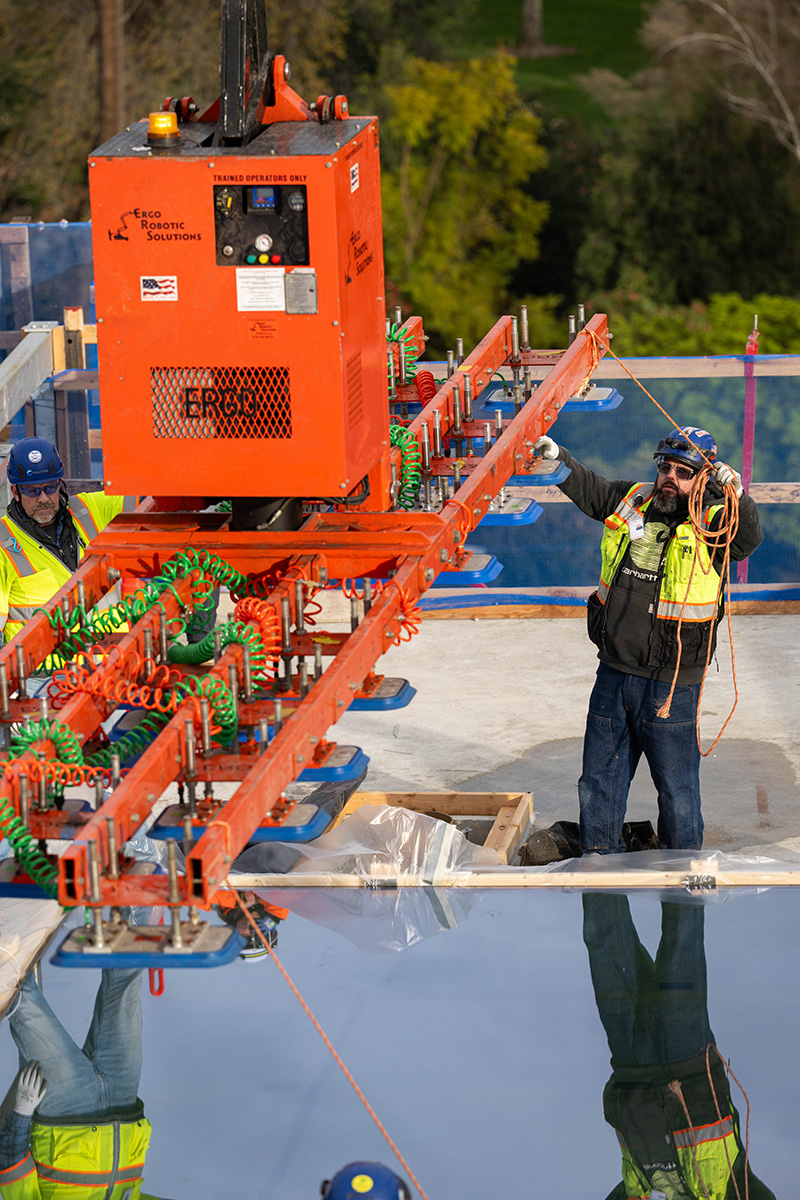 Construction workers wearing safety gear use a red robotic vacuum lifter to position a glass panel on the Capitol Annex roof.