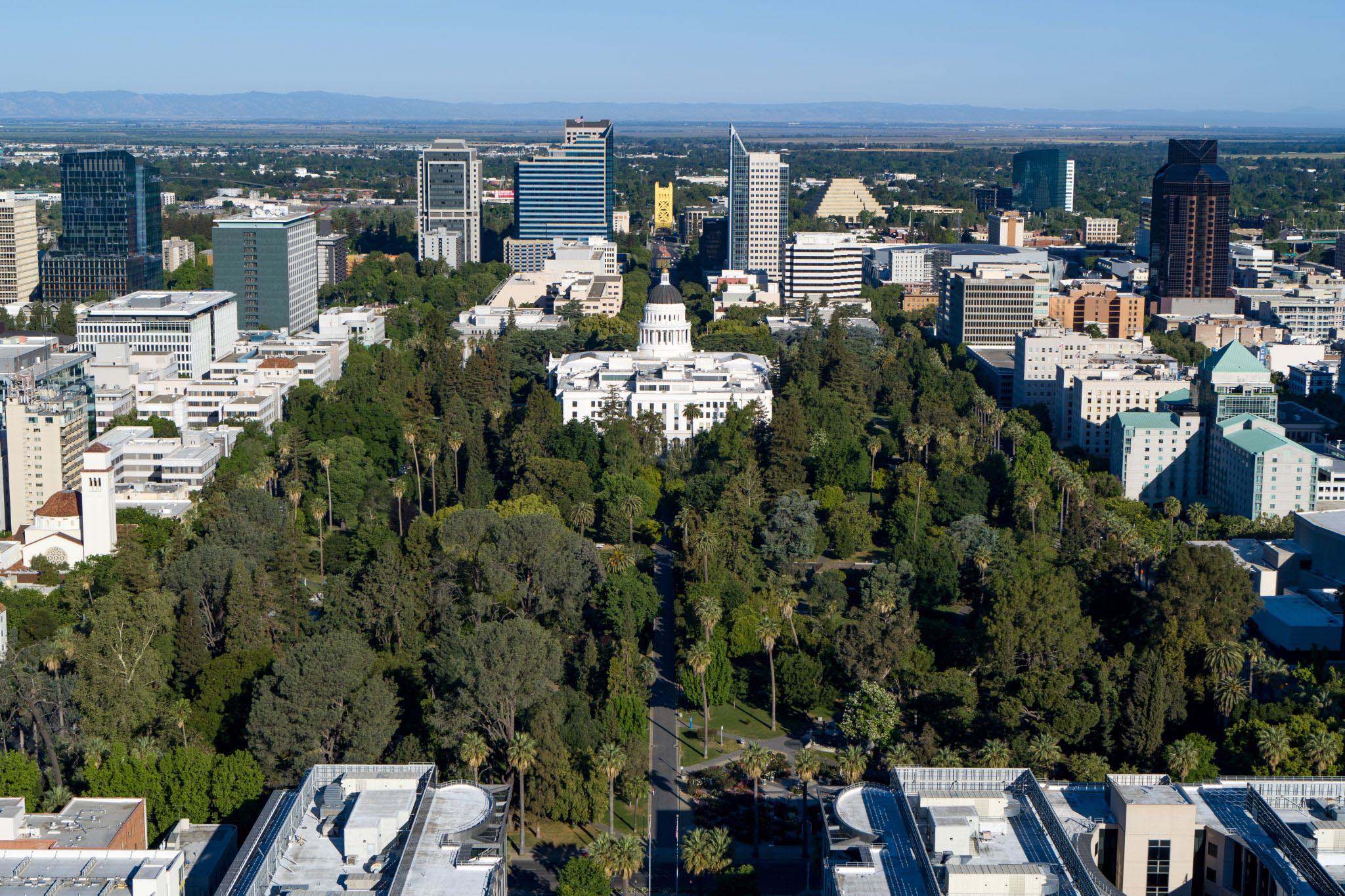 High-angle aerial view of downtown Sacramento, centering on the State Capitol building and the surrounding green trees of Capitol Park.