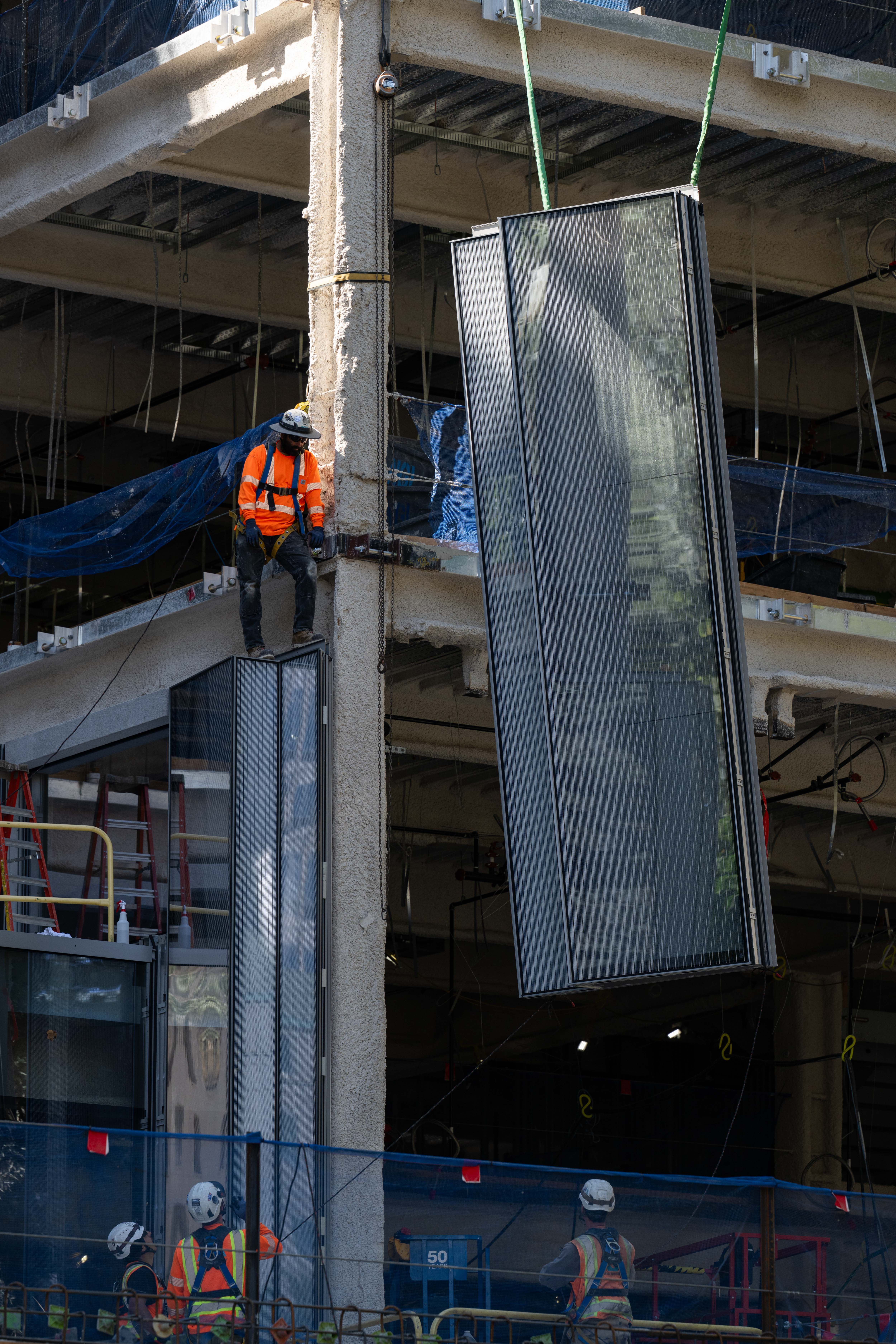 Construction crews look on as the crane operator places a façade glass panel into place.